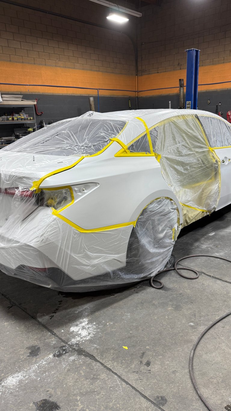 White car covered in protective plastic sheeting with yellow tape in an auto body shop garage