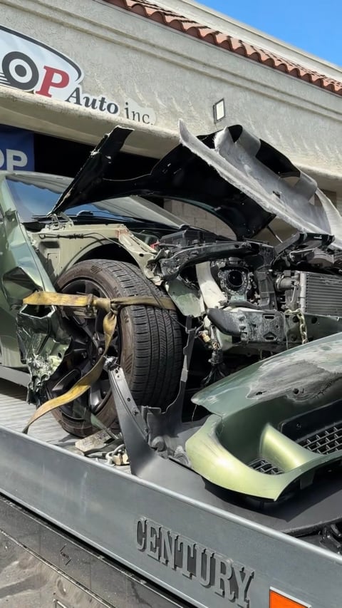 Severely damaged car with crushed hood and exposed engine being transported on a tow truck in front of an auto repair shop