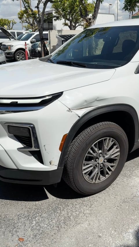 Close-up of white SUV front end showing hood, grille, and alloy wheel at dealership lot with trees and other vehicles in background