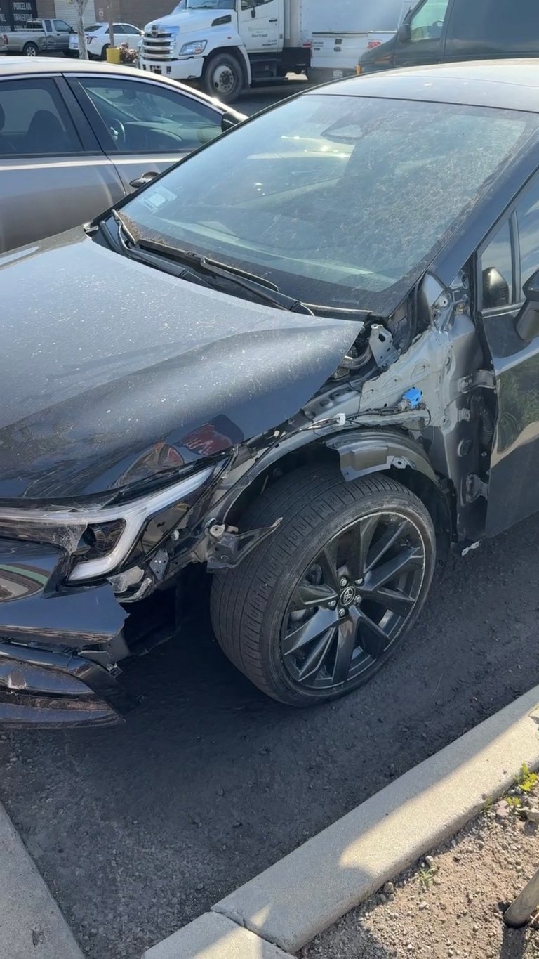 Damaged dark sedan with crumpled front end and hood in parking lot with other vehicles in background