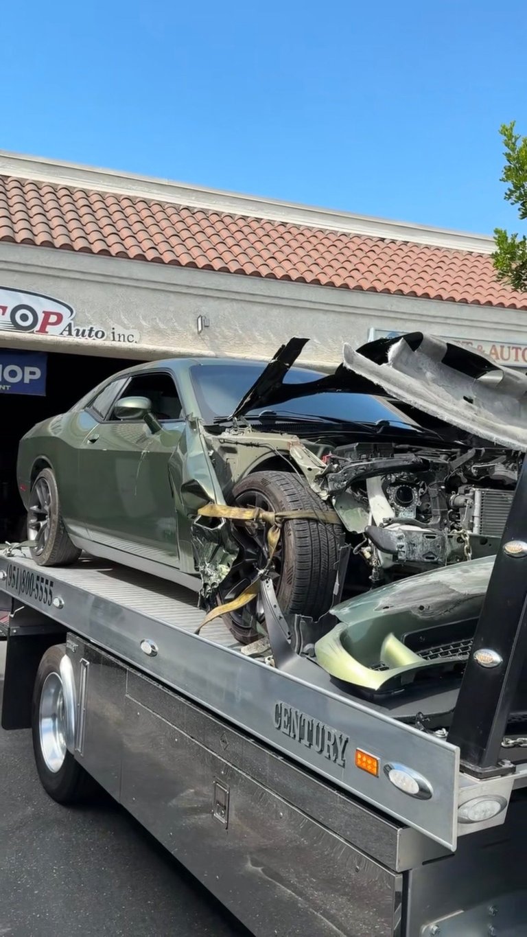 Severely damaged green car on a flatbed tow truck at an auto repair shop with clear sky and tiled building roof visible in background